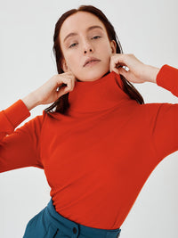 Close-up front view of a women's red merino wool hoodie by Šumska, with the woman holding the high, wide neck collar, highlighting the clean, minimal design and cozy fabric texture.