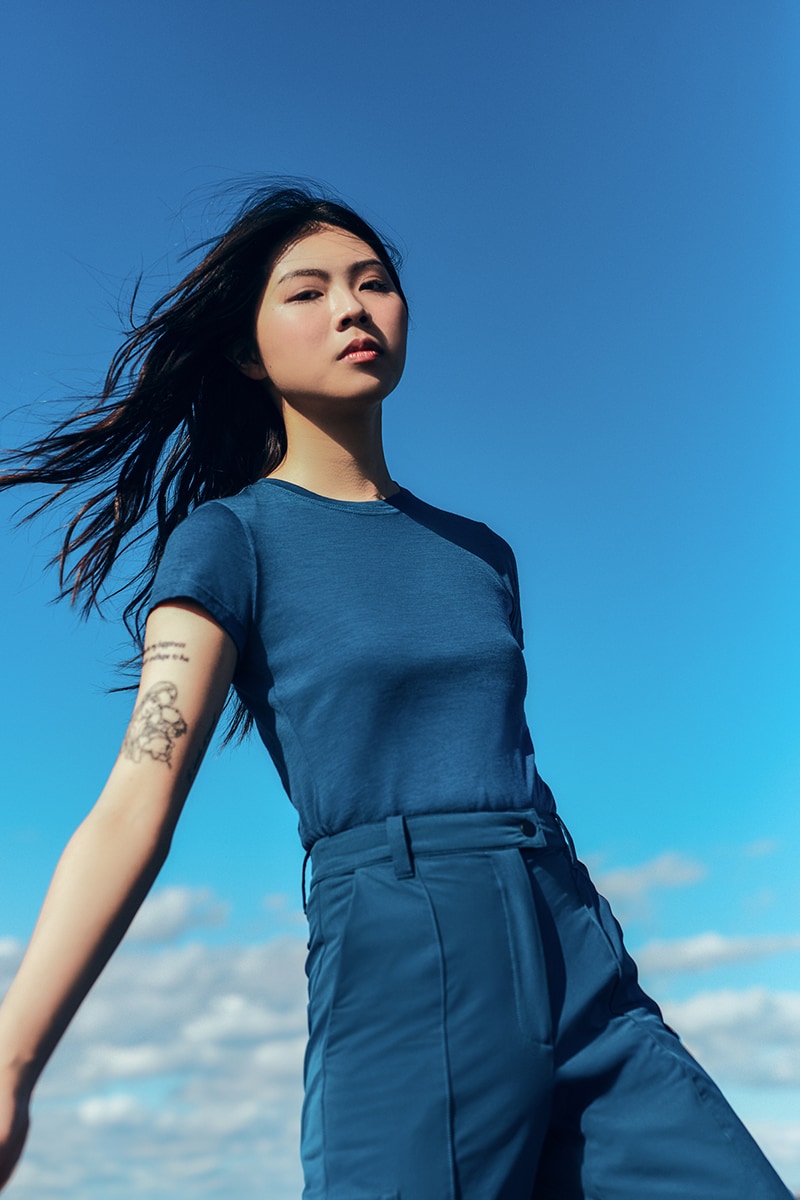 Model wearing the Šumska Blue Merino Classic T-Shirt paired with blue softshell hiking pants, standing outdoors with arms stretched out and windblown hair