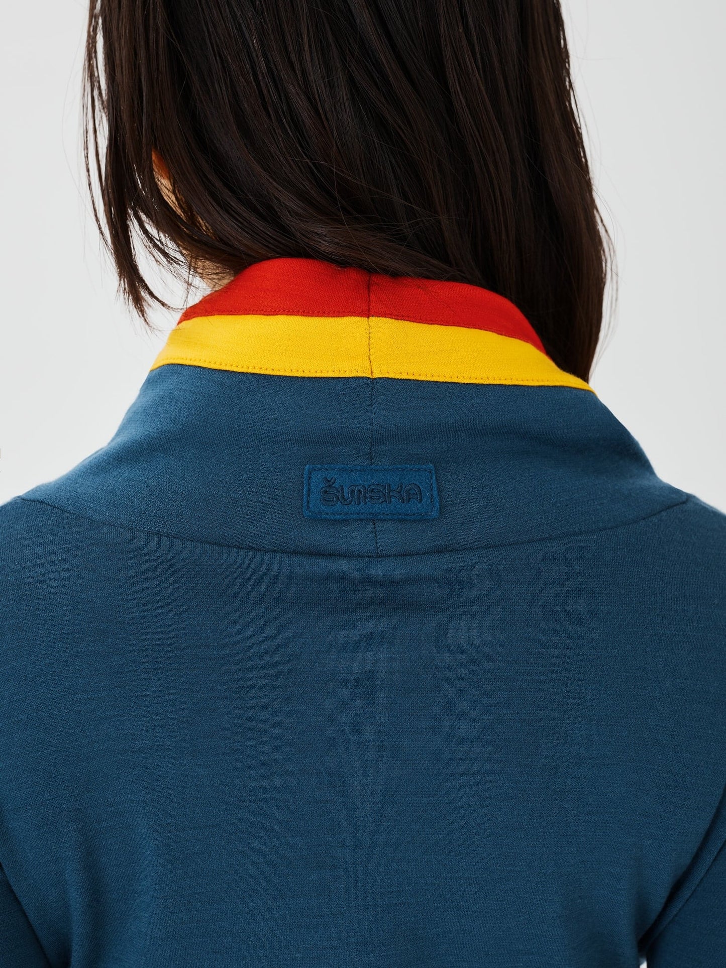 Close-up back view of a blue merino wool Twiggy pullover jumper by Šumska, showcasing the vibrant red and yellow stripe panels and an embroidered logo.