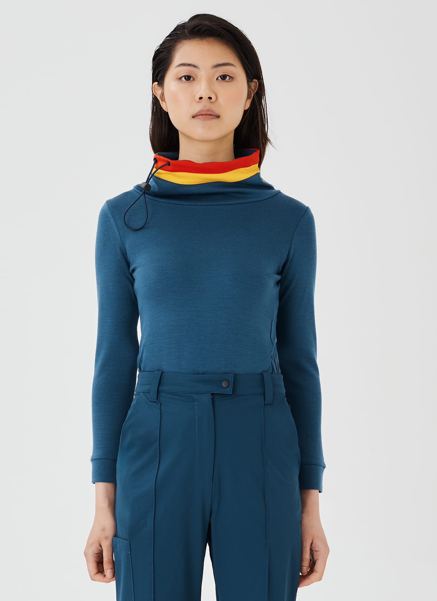 Close-up front view of a blue merino wool Twiggy pullover jumper by Šumska, featuring a wide collar with vibrant red and yellow stripe panels, paired with blue hiking pants