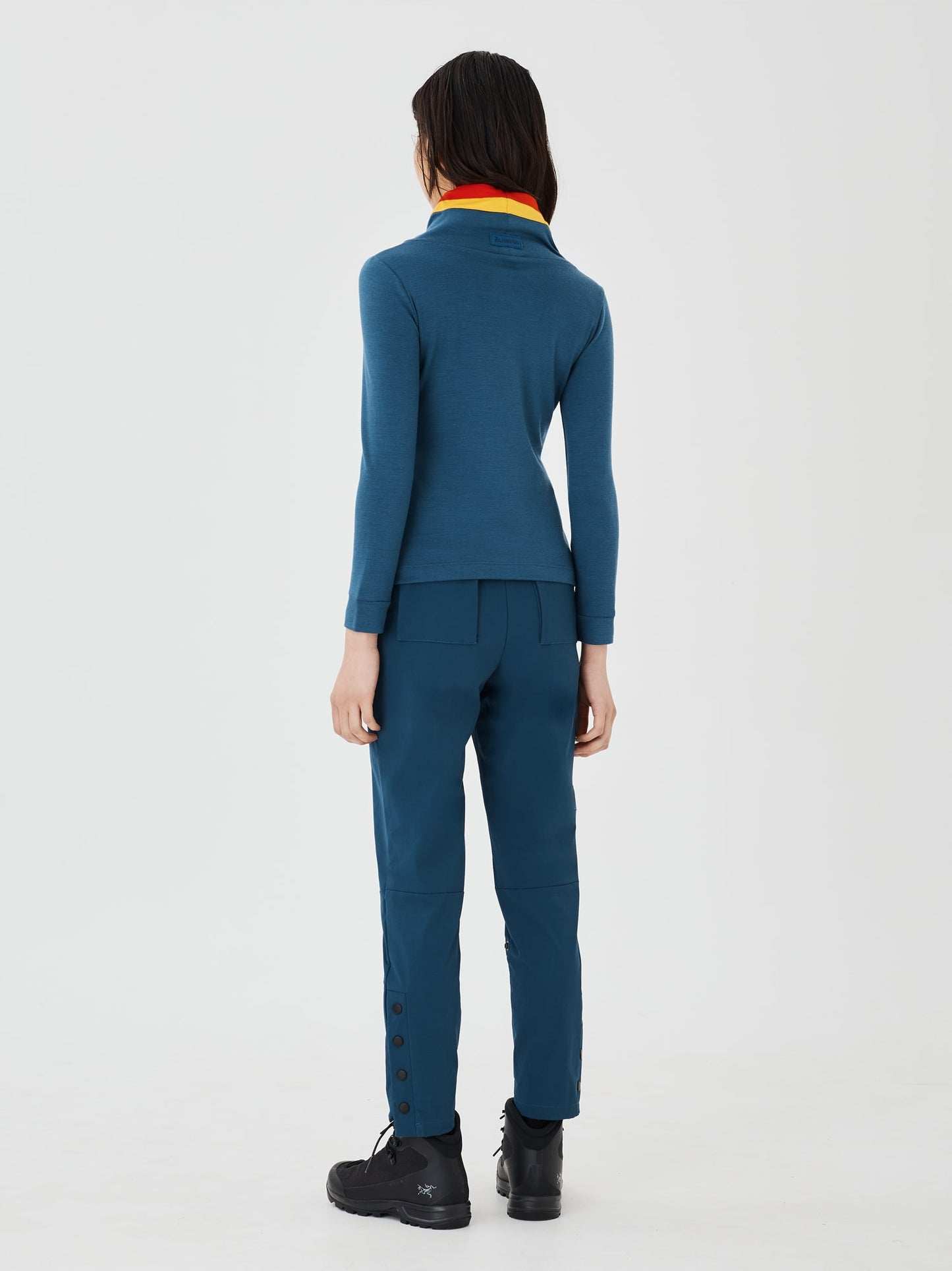 Full-body back view of a woman wearing a blue merino wool Twiggy pullover jumper by Šumska, paired with blue hiking pants, showcasing the vibrant red and yellow stripe panels.