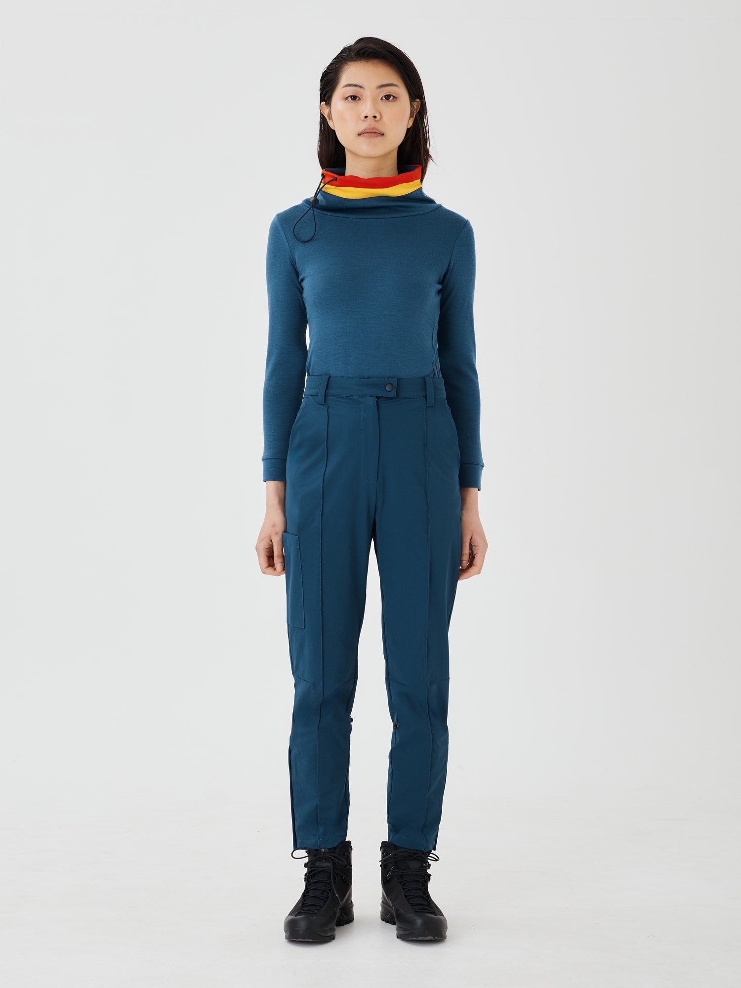 Full-body front view of a woman wearing a blue merino wool Twiggy pullover jumper by Šumska, featuring a wide collar with vibrant red and yellow stripe panels, paired with blue hiking pants.