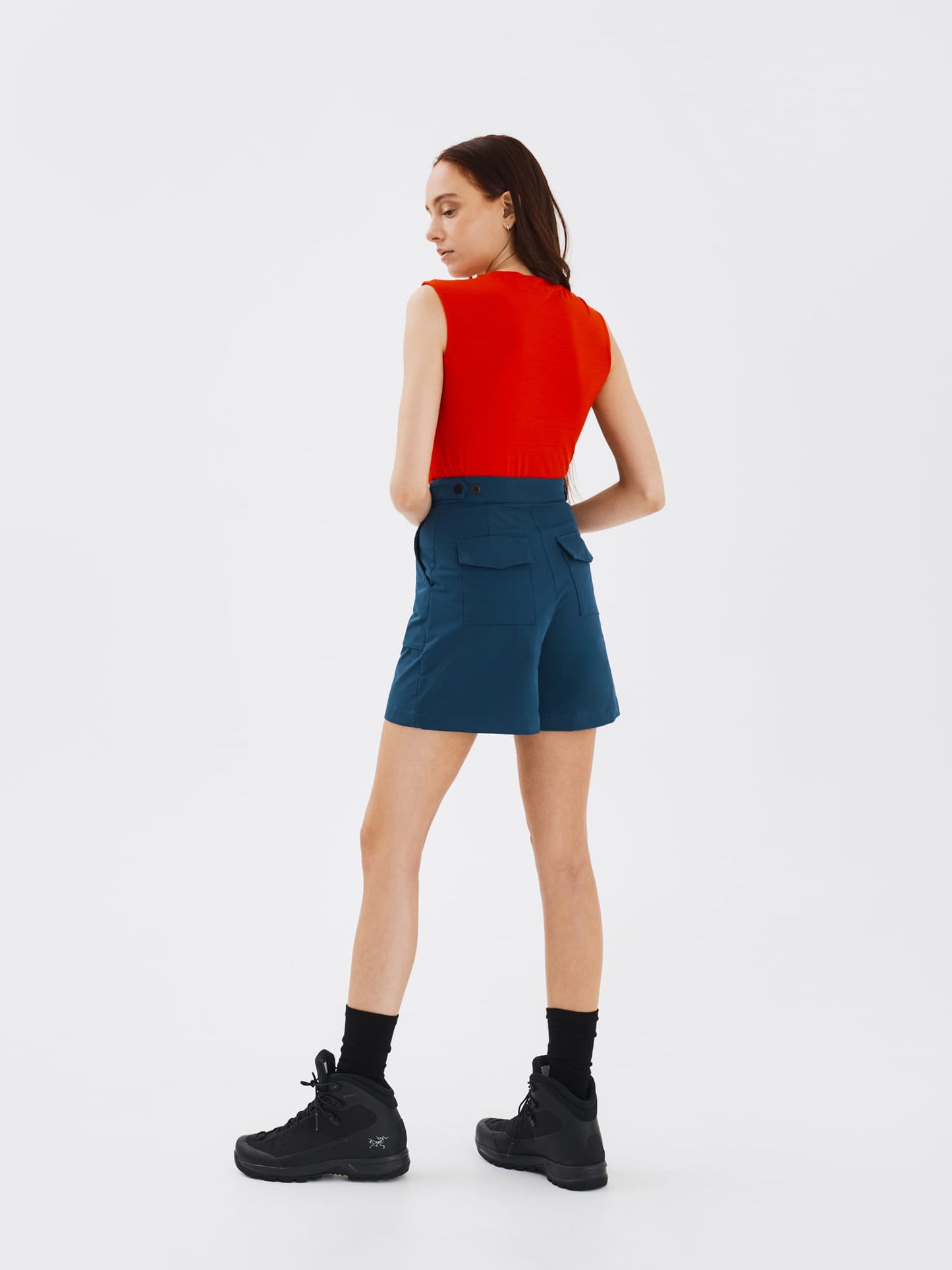 Full-body back view of a woman wearing blue Softshell Hiking Shorts by Šumska, paired with a red merino sleeveless T-shirt, showcasing a classic outdoor style and comfortable fit.