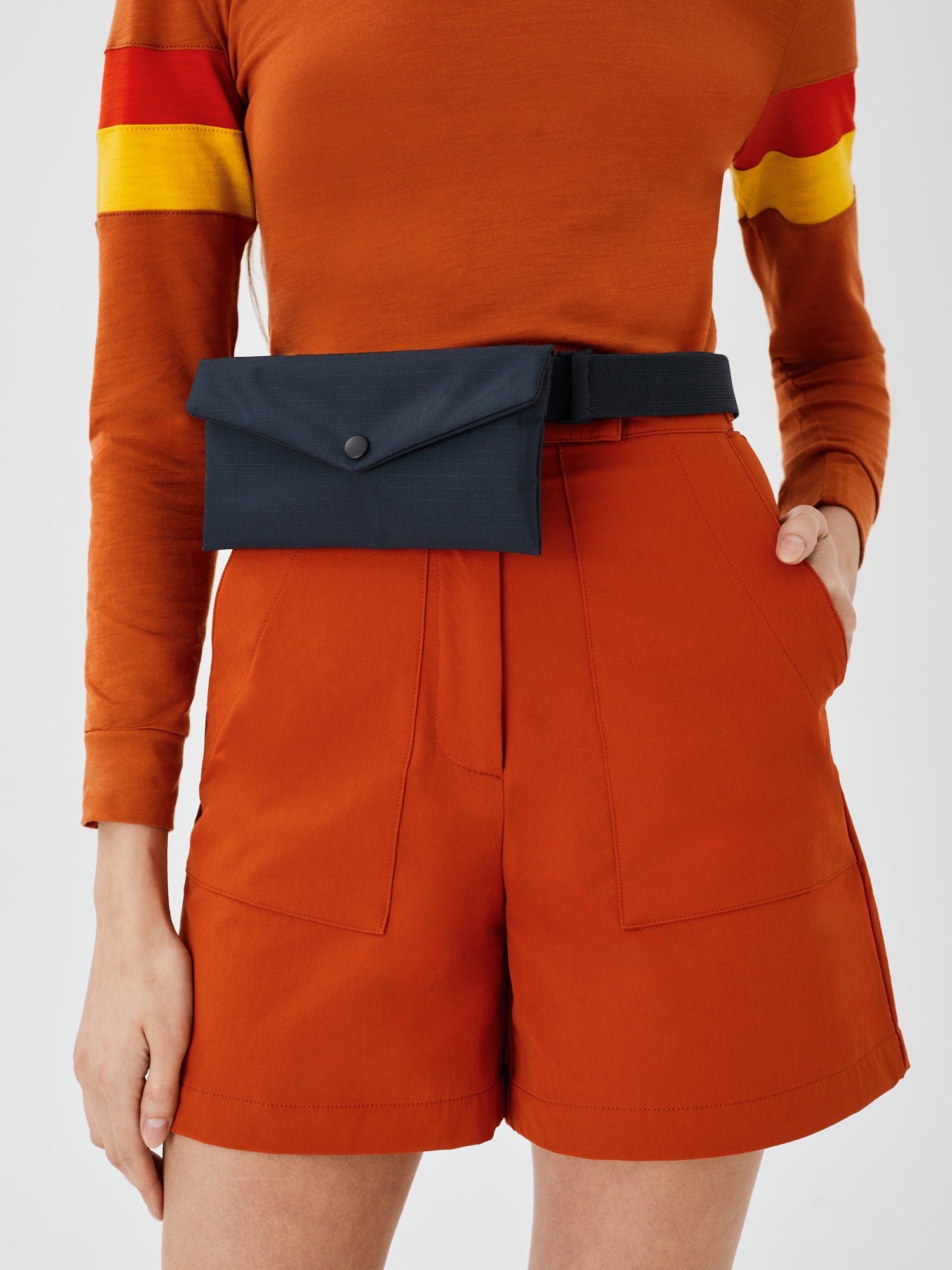 Close-up front view of a midnight blue Cordura Belt Bag by Šumska on a woman's waist, wearing rust hiking softshell shorts and a long-sleeve rust merino top, highlighting the compact design and durable fabric.