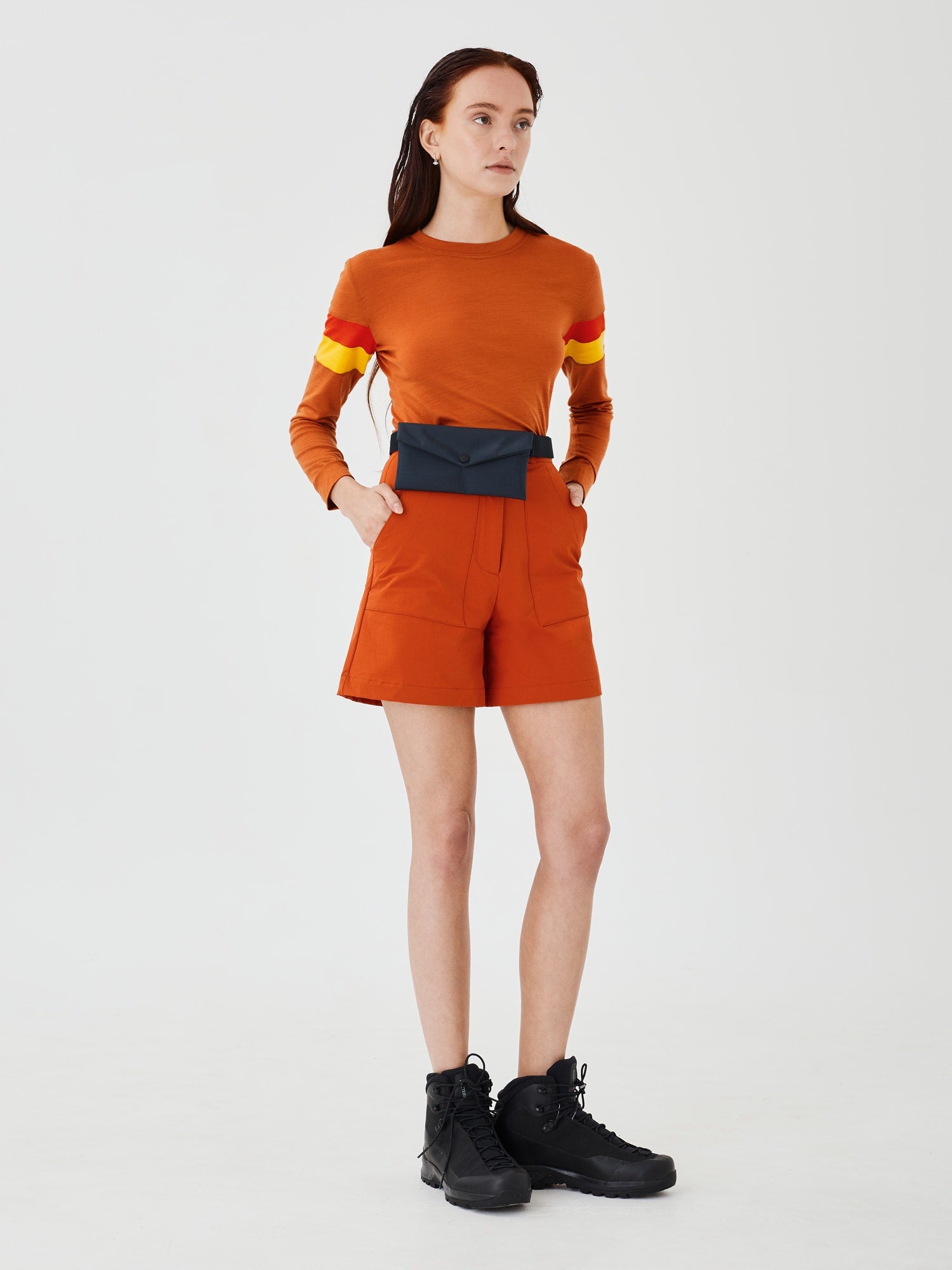 Full body front view of a woman wearing a midnight blue Cordura Belt Bag by Šumska, paired with rust hiking softshell shorts and a long-sleeve rust merino top, highlighting the bag's compact design and durable fabric."