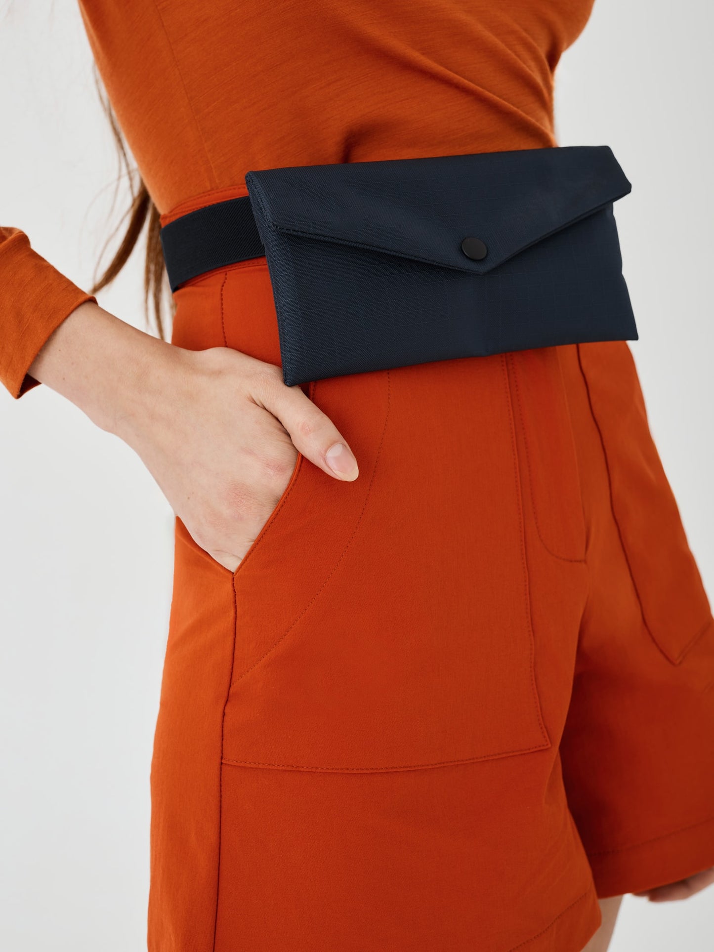 Zoomed-in view of a midnight blue Cordura Belt Bag by Šumska, attached with a black belt at the waist of a woman wearing rust hiking softshell shorts and a long-sleeve rust merino top."