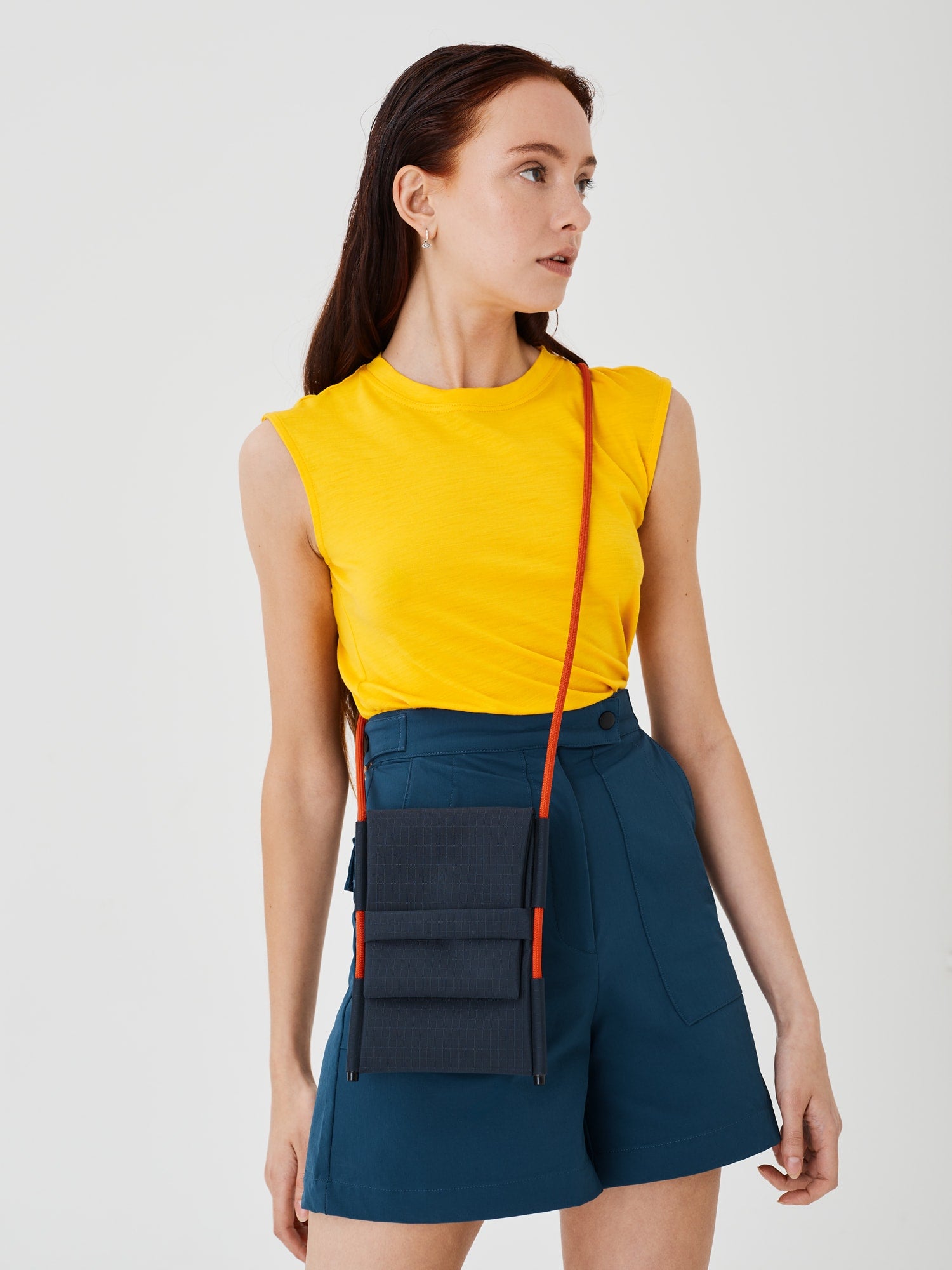 Close-up angled view of a woman wearing the Šumska Cordura Crossbody iPhone Bag with a solar orange Paracord strap, paired with blue softshell hiking shorts and a yellow sleeveless T-shirt.