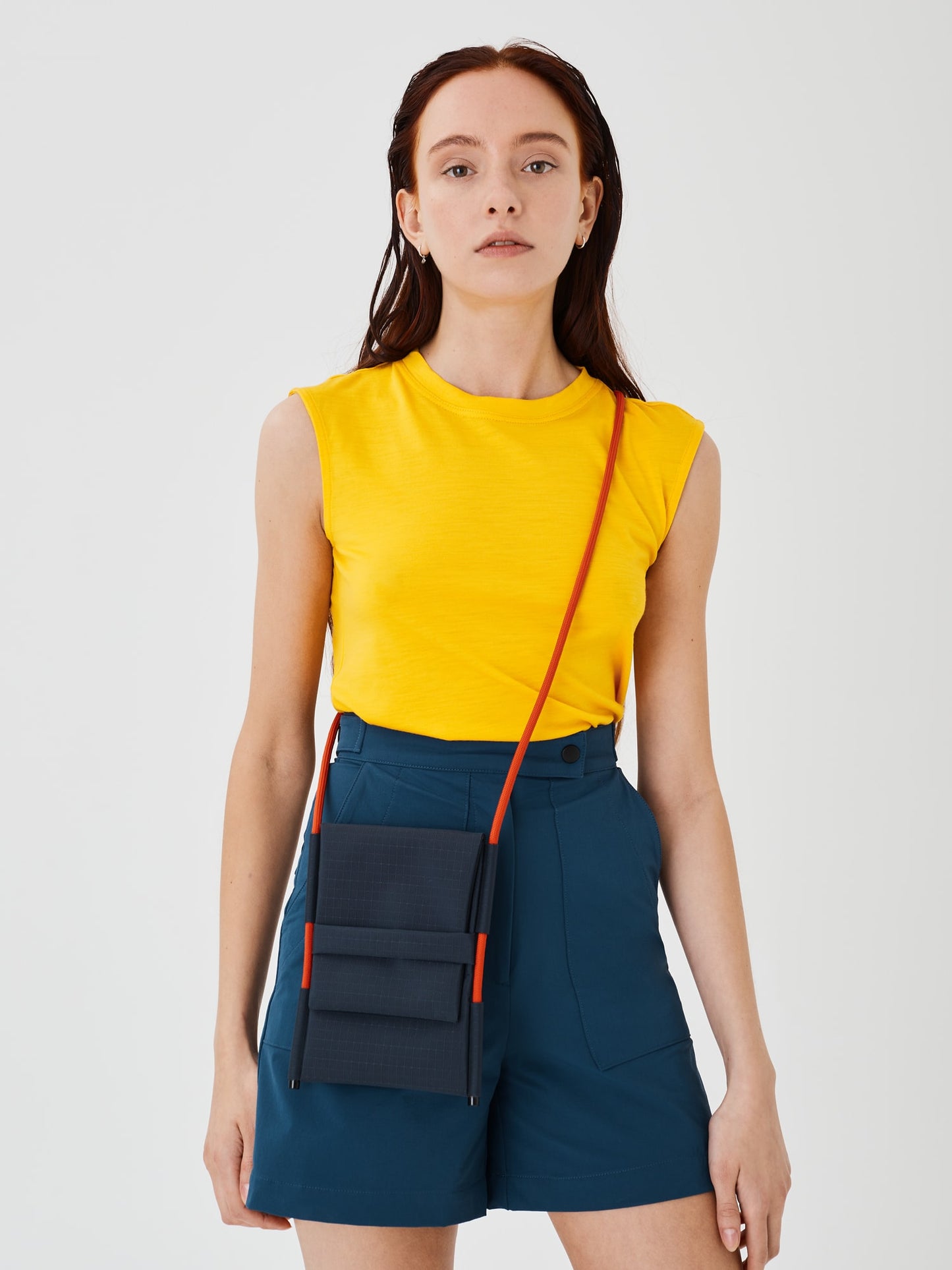 Close-up straight view of a woman wearing the Šumska Cordura Crossbody iPhone Bag with a solar orange Paracord strap, paired with blue softshell hiking shorts and a yellow sleeveless T-shirt.