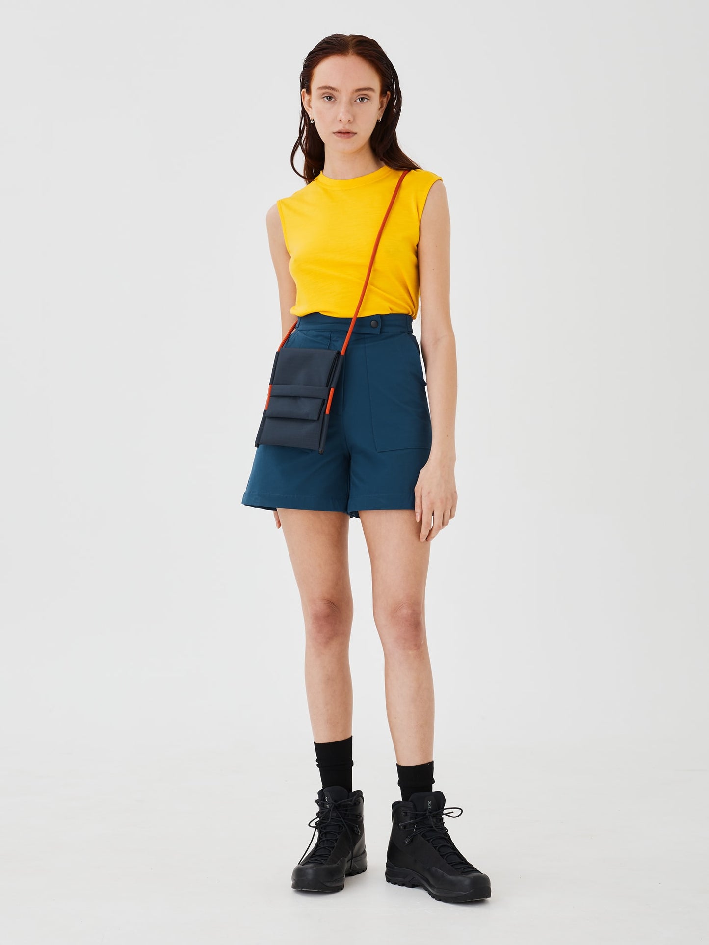 Full-body front view of a woman wearing the Šumska Cordura Crossbody iPhone Bag with a solar orange Paracord strap, paired with blue softshell hiking shorts and a yellow sleeveless T-shirt.