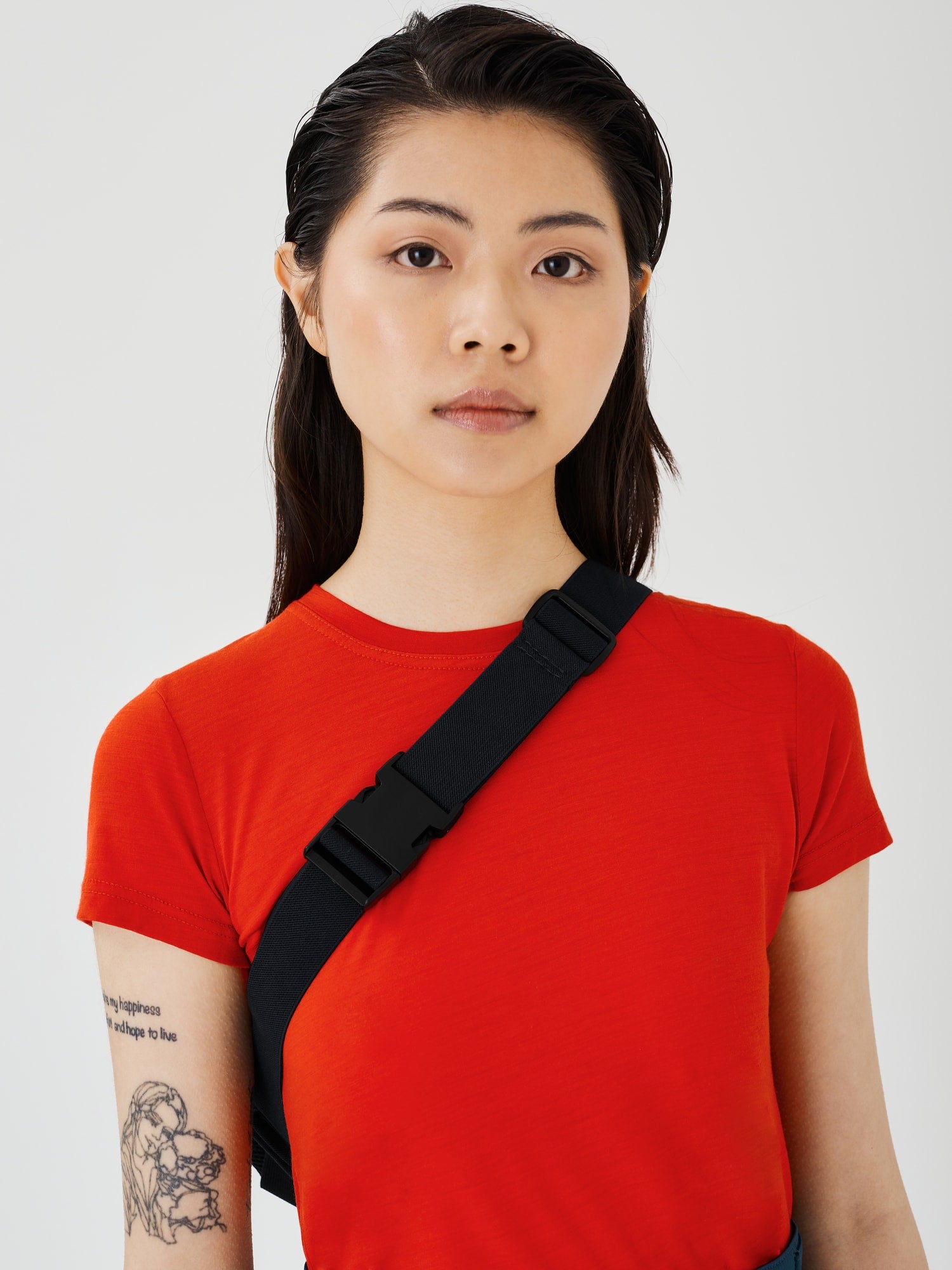 View of a woman wearing a red merino classic T-shirt, showing the black elastic strap of the Šumska midnight blue Cordura sling bag across the body, with a black plastic buckle and adjustable glider.