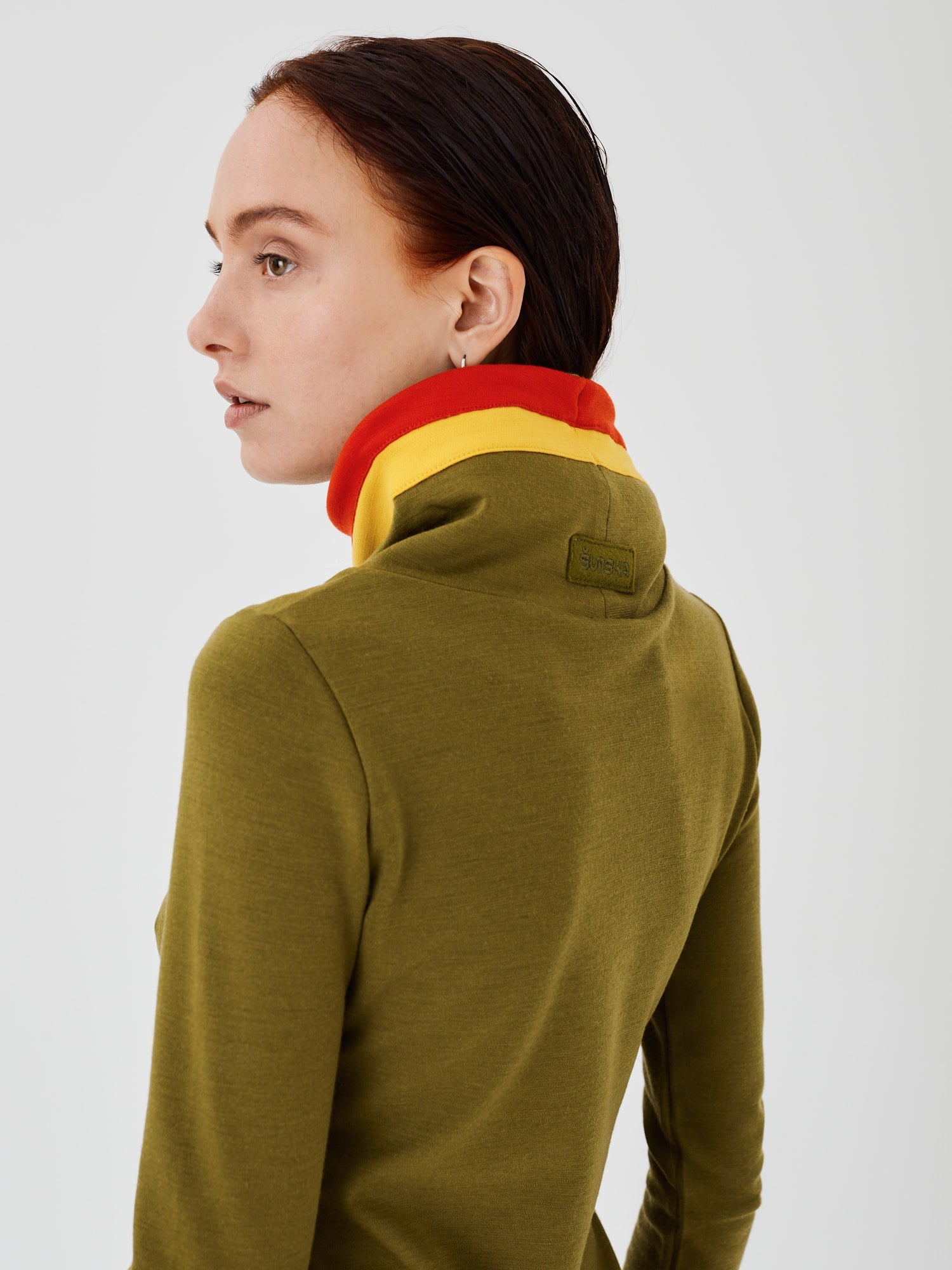 Close-up back view of an olive green merino wool Twiggy pullover jumper by Šumska, showcasing the embroidered logo and vibrant red and yellow stripe panels.