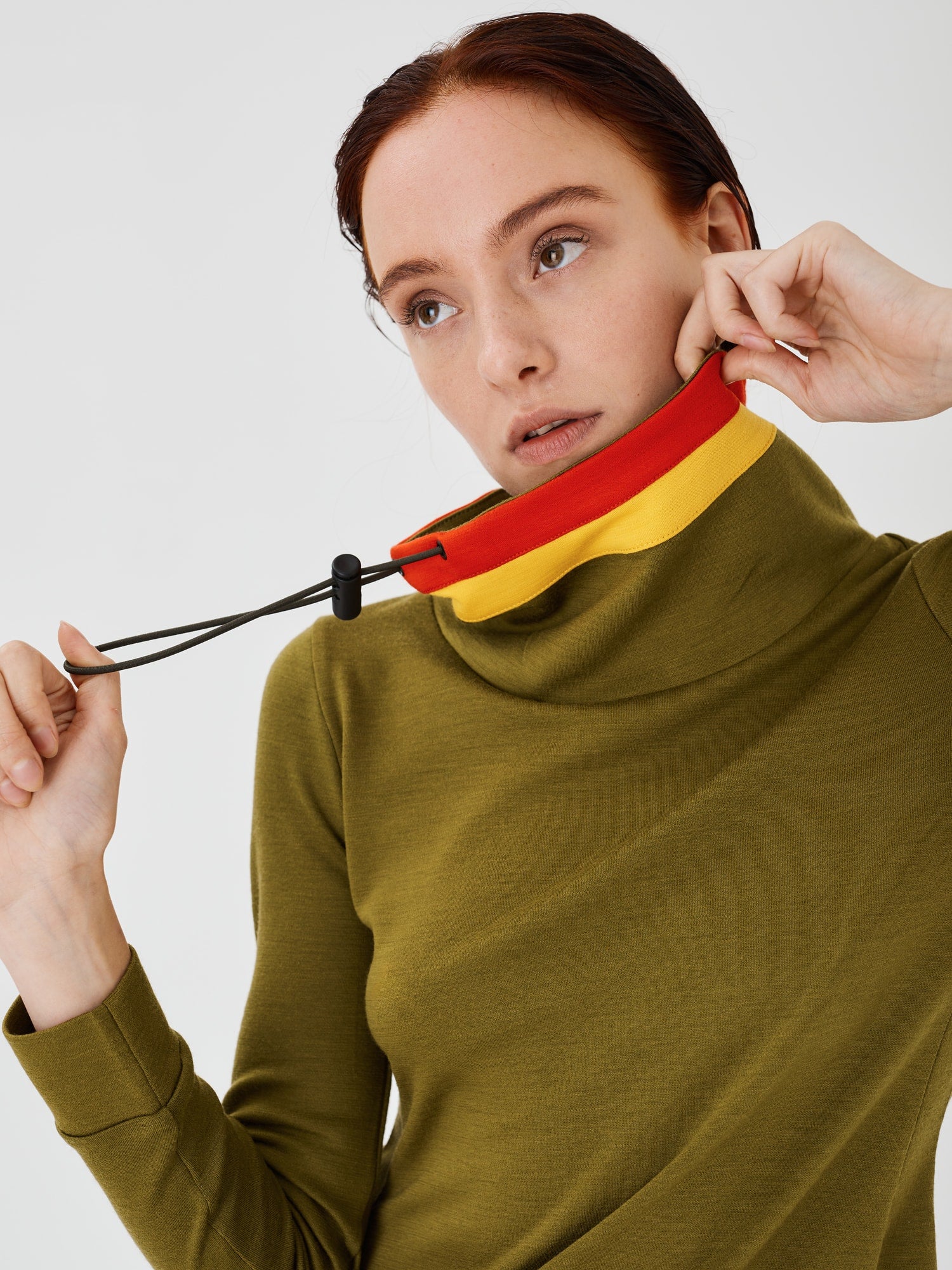 Close-up view of a woman holding the collar of an olive green merino wool Twiggy pullover jumper by Šumska with one hand, while pulling the drawcord with the other, featuring vibrant red and yellow stripe panels.