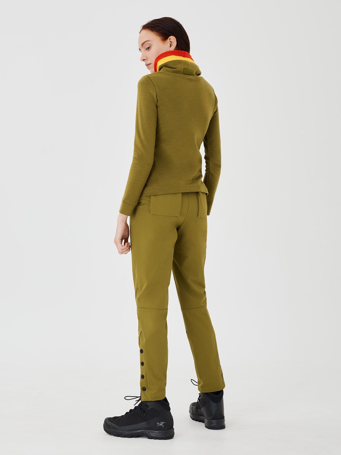 Full-body back view of a woman wearing an olive green merino wool Twiggy pullover jumper by Šumska, featuring vibrant red and yellow stripe panels, paired with olive green hiking pants.