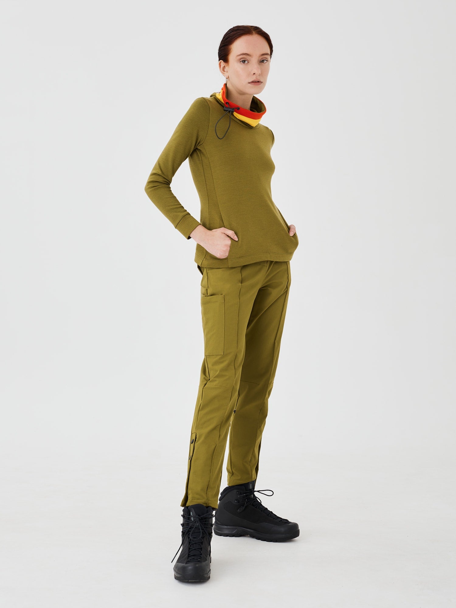 Full front side view of a woman wearing an olive green merino wool Twiggy pullover jumper by Šumska, with her hand in the pockets, featuring vibrant red and yellow stripe panels, paired with olive green hiking pants.