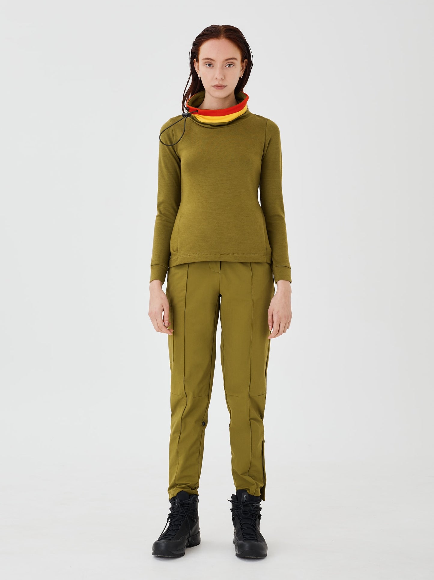 Full front view of a woman wearing an olive green merino wool Twiggy pullover jumper by Šumska, paired with olive green hiking pants, featuring a stylish design with vibrant red and yellow stripe panels.