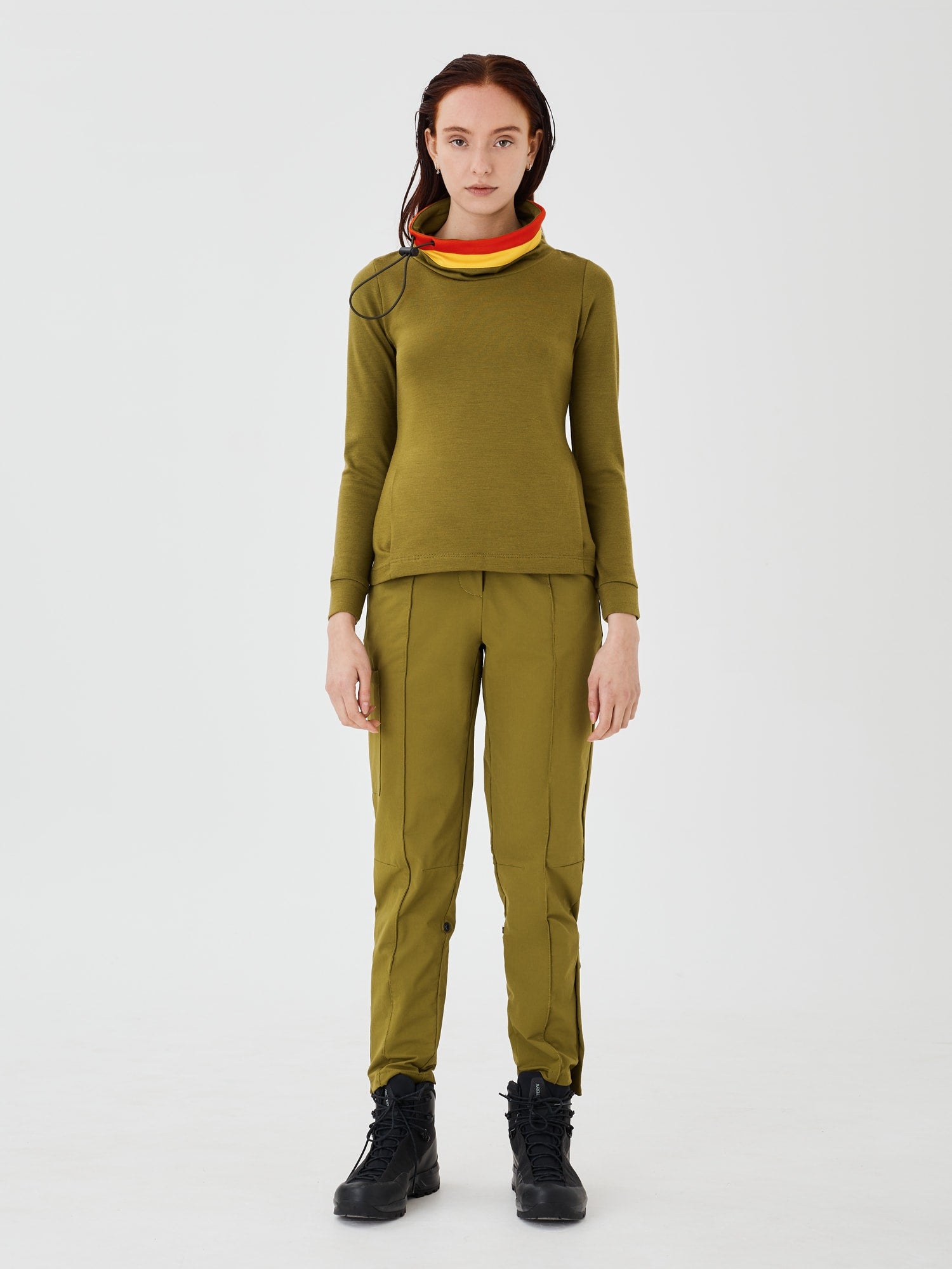 Full front view of a woman wearing an olive green merino wool Twiggy pullover jumper by Šumska, paired with olive green hiking pants, featuring a stylish design with vibrant red and yellow stripe panels.