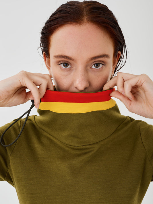 Zoomed-in view of a woman holding the collar of an olive green merino wool Twiggy pullover jumper by Šumska, featuring vibrant red and yellow stripe panels.