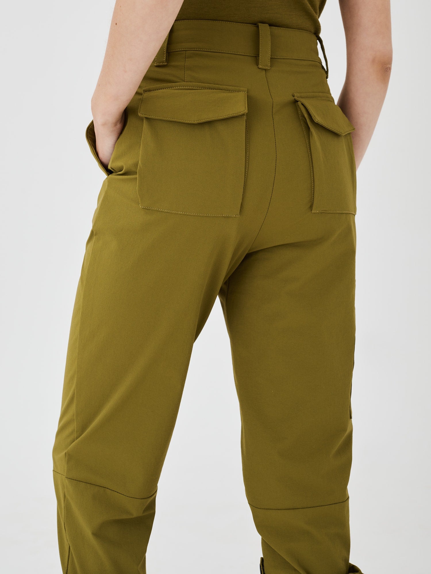 Close-up back view of olive green Softshell Hiking Pants by Šumska, showing a woman with a hand in the front pocket, back pockets, belt loops, and articulated knee feature.