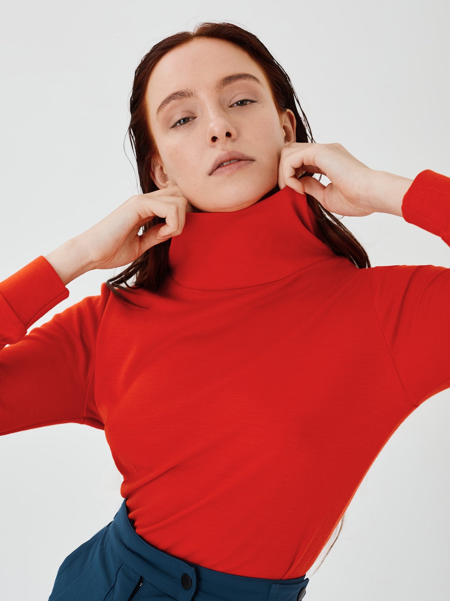 Close-up front view of a women's red merino wool hoodie by Šumska, with the woman holding the high, wide neck collar, highlighting the clean, minimal design and cozy fabric texture.