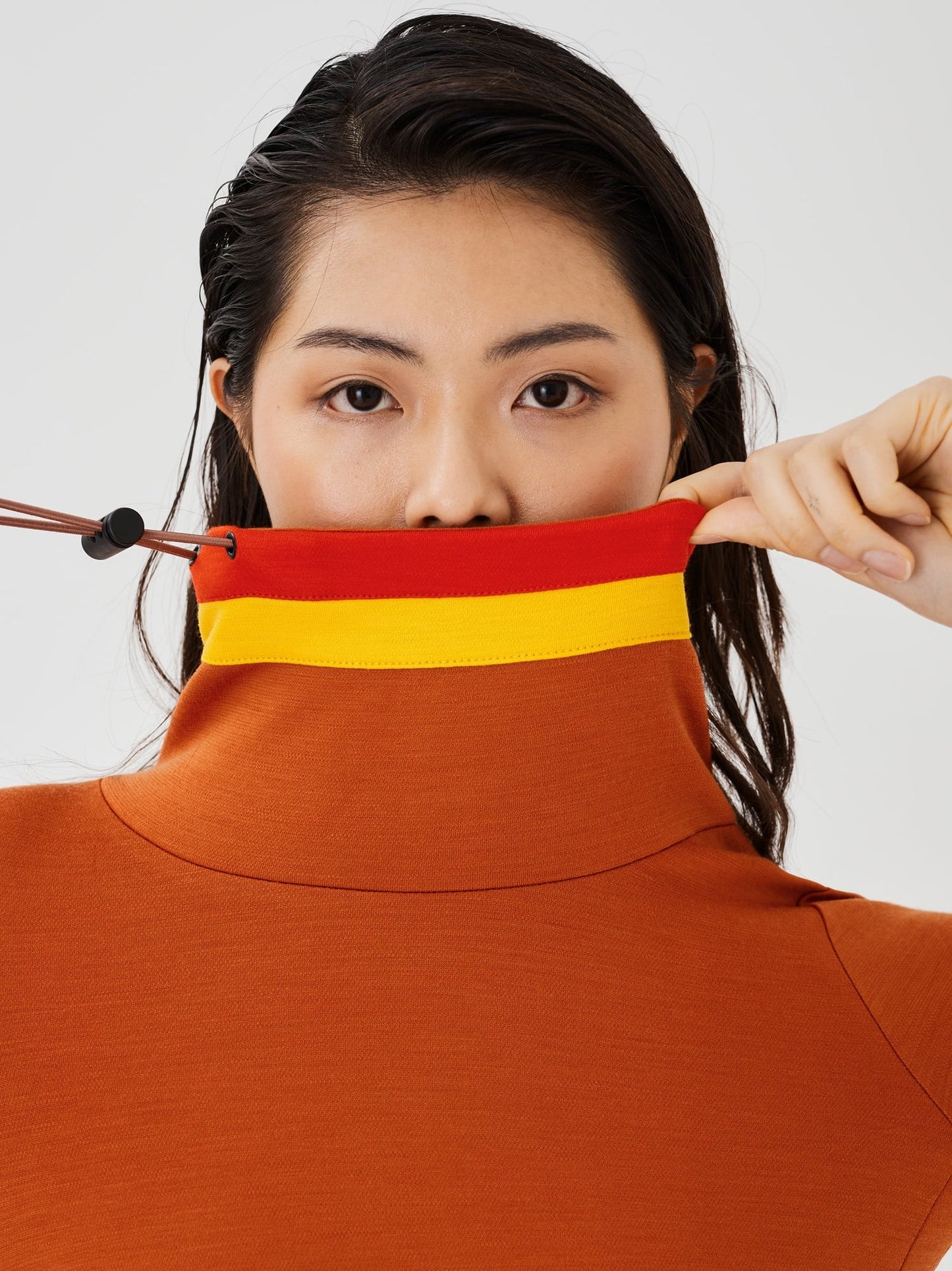 Closer zoomed-in view of a woman holding the drawstring and collar of a rust-coloured merino wool Twiggy pullover jumper by Šumska, highlighting the vibrant red and yellow stripe panels.