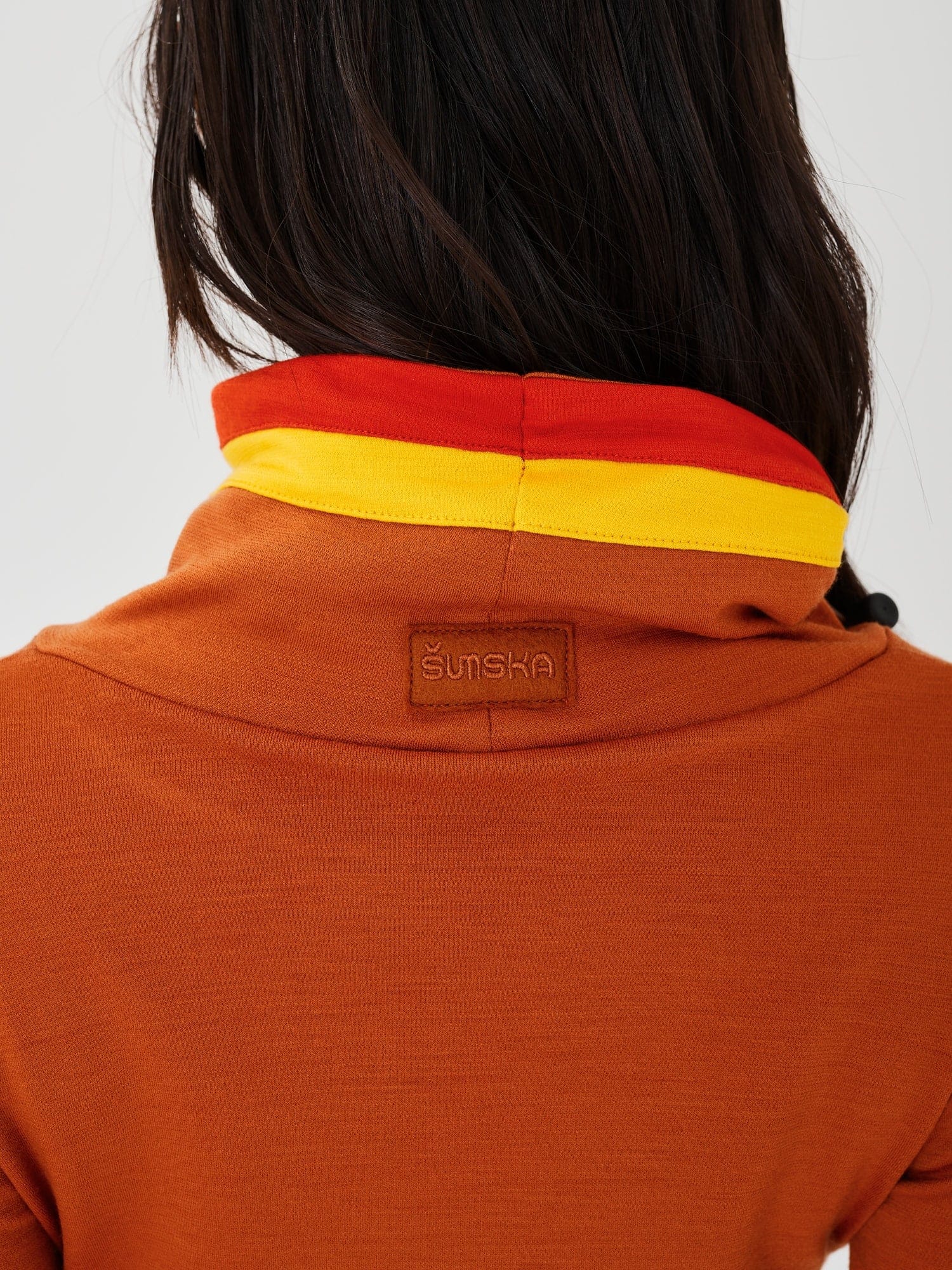 Close-up back view of a rust-colored merino wool Twiggy pullover jumper by Šumska, showcasing the embroidered logo and vibrant red and yellow stripe panels.