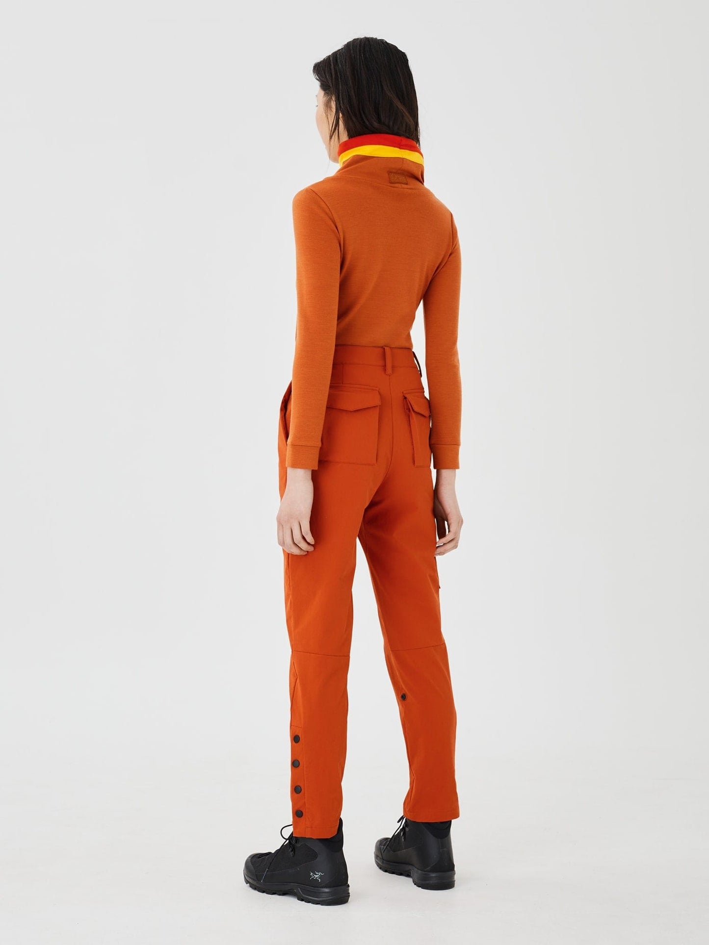 Full-body back view of a woman wearing a rust-colored merino wool Twiggy pullover jumper by Šumska, featuring vibrant red and yellow stripe panels, paired with rust hiking pants.