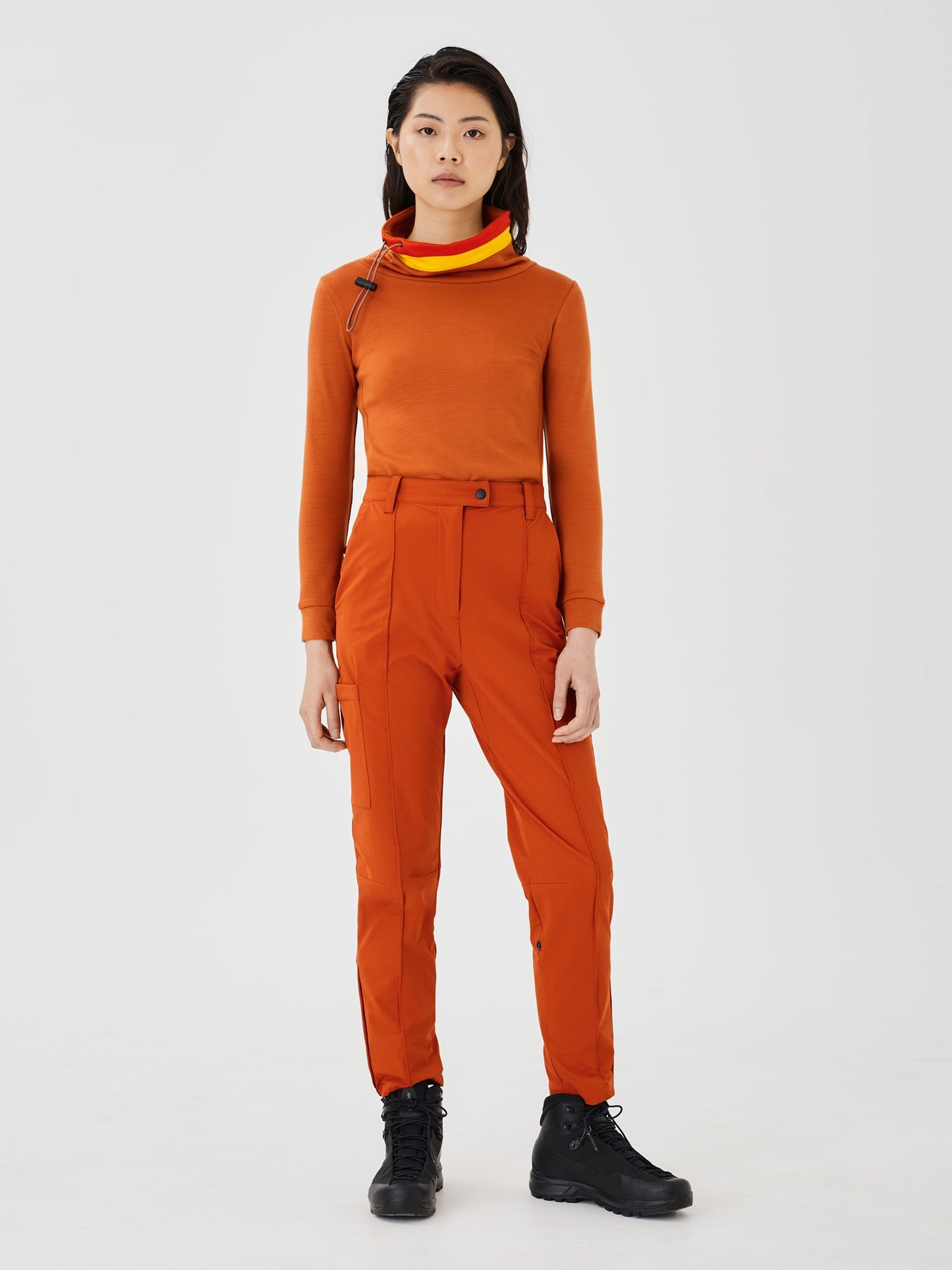 Full-body front view of a woman wearing a rust-colored merino wool Twiggy pullover jumper by Šumska, featuring vibrant red and yellow stripe panels, paired with rust hiking pants.