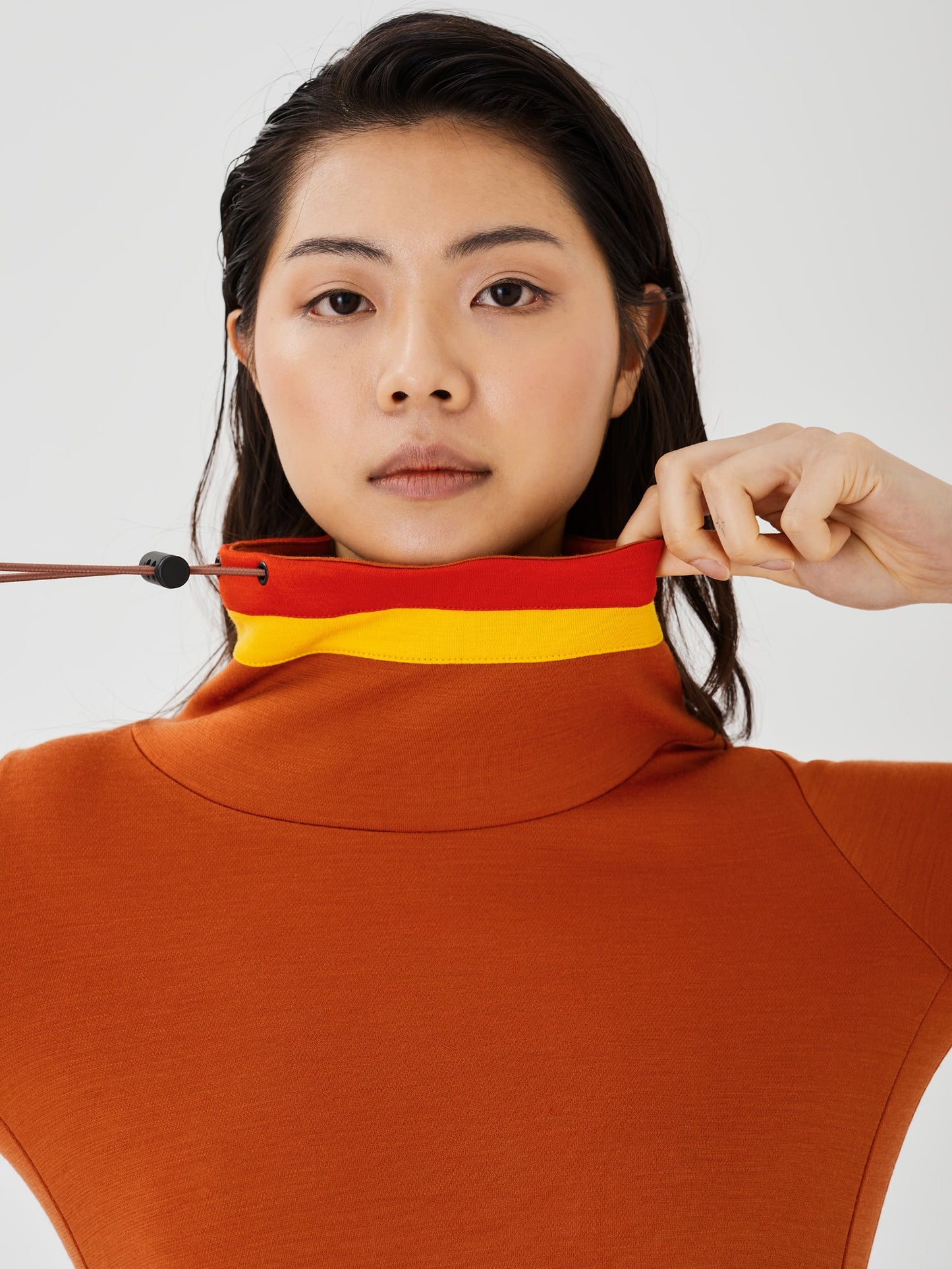 Zoomed-in view of a woman holding the drawstring and collar of a rust-coloured merino wool Twiggy pullover jumper by Šumska, featuring vibrant red and yellow stripe panels.