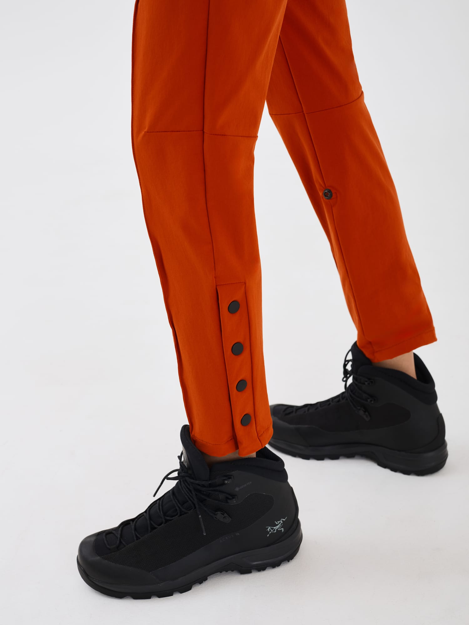 Close-up view of the ankle gusset on rust-coloured Softshell Hiking Pants by Šumska, highlighting the black snap button features, versatile design, and articulated knee detail.