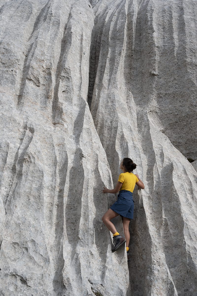 Image of woman free climbing on white granite rock wall wearing yellow merino Classic T-shirt and blue Softshell Shorts by Šumska