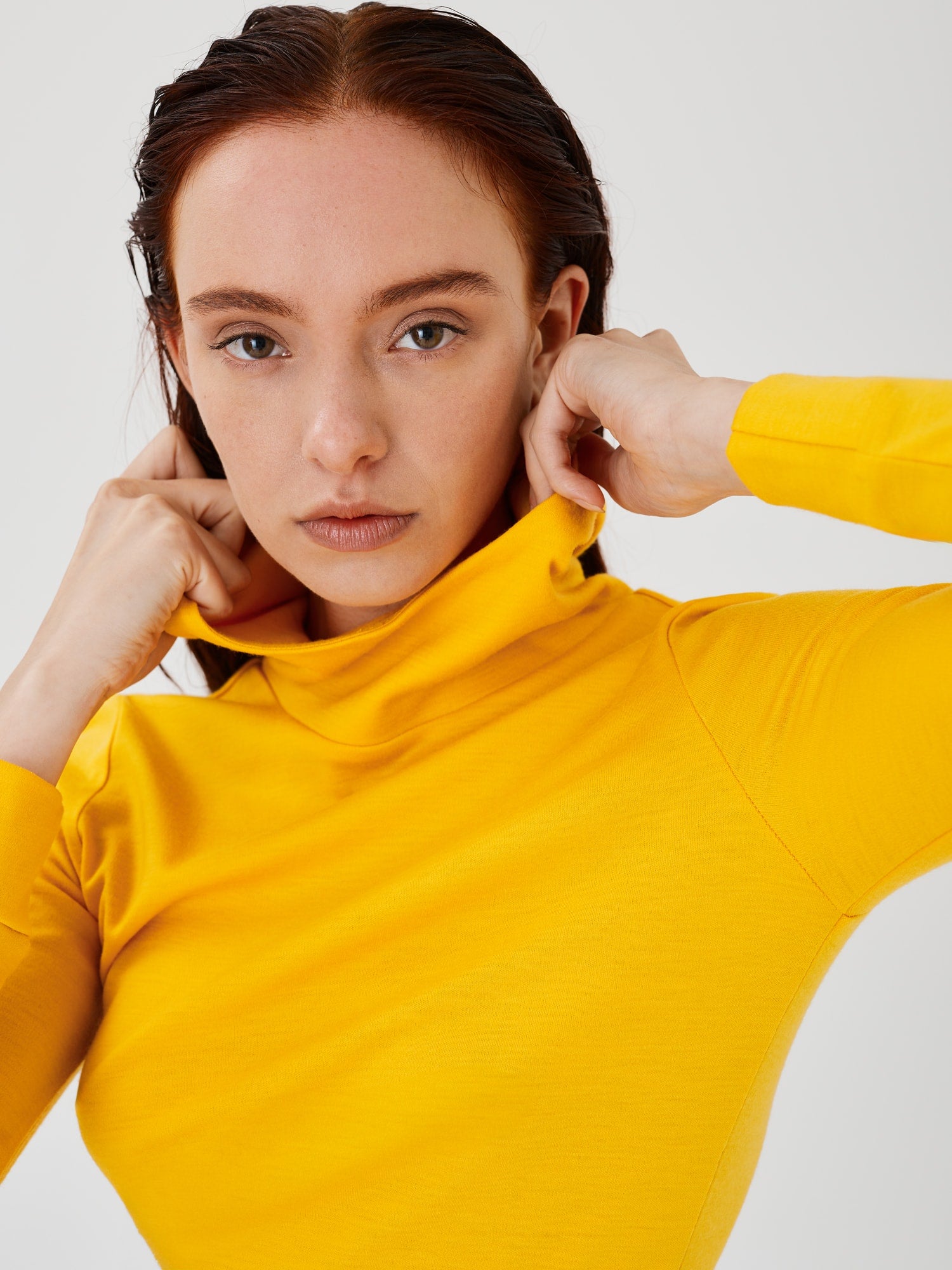 Close-up front view of a women's yellow merino wool hoodie by Šumska, with the woman holding the collar, highlighting the clean, minimal design and soft, cozy fabric.